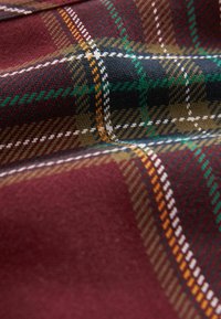 Tartan stof i nuancer af bordeaux, grøn, sort og brun med en struktureret vævning og kontrasterende hvide og orange linjer.