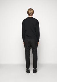 Mann mit hellbraunen Haaren, der einen schwarzen Pullover, schwarze Hosen, weiße Socken und schwarze Schuhe trägt und vor einer leeren weißen Wand steht.