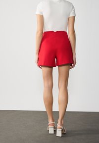 Czerwone szorty z wysokim stanem o dopasowanym kroju, z bocznymi kieszeniami i gładkim wykończeniem. W połączeniu z białym topem z krótkim rękawem oraz szarymi butami na słupku.