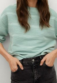 Femme aux longs cheveux bruns portant un pull côtelé bleu clair et un jean noir taille haute, les mains dans les poches.