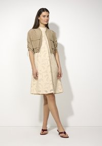 Hellbeiges Kleid mit strukturiertem Blumenmuster und leicht ausgestelltem Saum, kombiniert mit einer taupefarbenen cropped Jacke und burgunderfarbenen Sandalen.