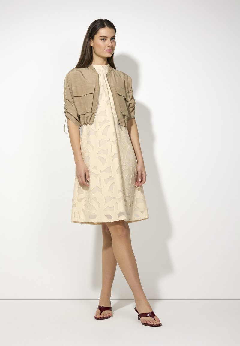 Hellbeiges Kleid mit strukturiertem Blumenmuster und leicht ausgestelltem Saum, kombiniert mit einer taupefarbenen cropped Jacke und burgunderfarbenen Sandalen.