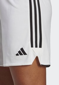Weiße Sportshorts mit schwarzen Seitenstreifen, einem belüfteten Saum und einem schwarzen gestickten Logo am unteren linken Rand. Glatte Stofftextur.