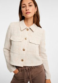 Crème geklede jasje met een kraag, voorzien van twee voorzakken, gouden knopen en textielstof. Draag het met een paar bruine broeken.