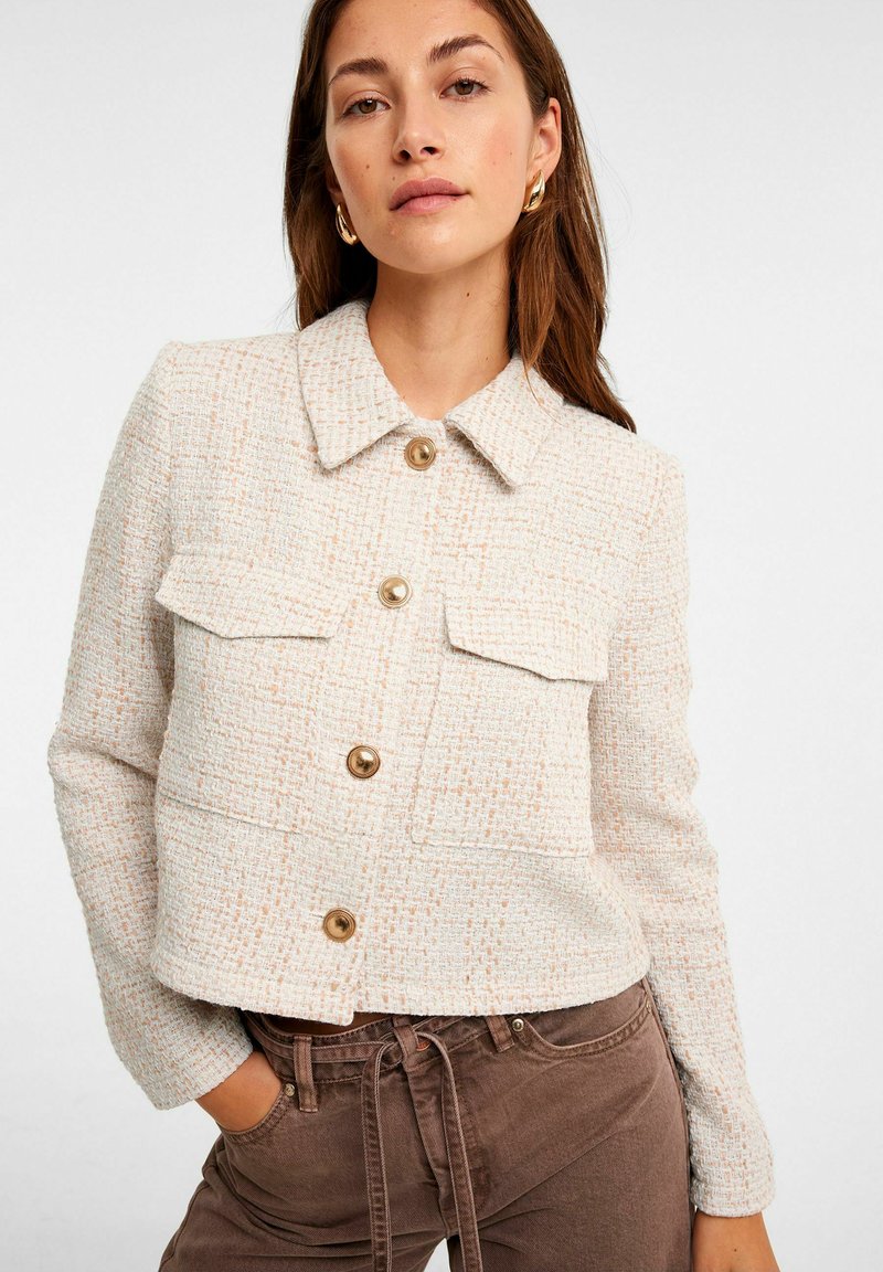 Crème geklede jasje met een kraag, voorzien van twee voorzakken, gouden knopen en textielstof. Draag het met een paar bruine broeken.