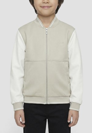 Niño con chaqueta beige y blanca con cremallera y pantalones negros, de pie frente a un fondo claro y liso.