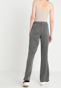 Kvinna iklädd grå högmidjade vida byxor, beigemönstrad ärmlös ribbad topp med djup rygg, och vita sneakers, stående med ryggen vänd.