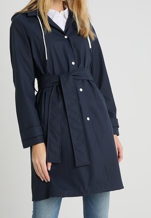 Femme portant un trench-coat bleu marine ceinturé avec des boutons-pression blancs sur une chemise blanche et un jean bleu.