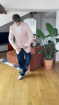 Mladic zakopčava svijetlu ružičastu jaknu preko bijele košulje, u tamnim trapericama i srebrnim cipelama, stojeći u modernom dnevnom boravku s velikom biljkom.