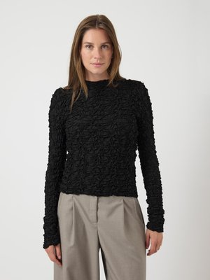 Black long-sleeve top with textured, gathered fabric. The design features a high neck and form-fitting silhouette, paired with light gray trousers.