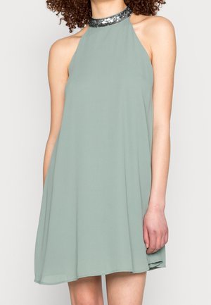 Robe à col halter en tissu vert doux avec une silhouette évasée. Présente un col noir pailleté. Texture lisse, longueur genoux.