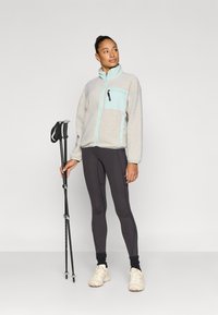 Ζακέτα φλις σε ανοιχτό γκρι με τσέπη στο στήθος σε μέντα, συνδυασμένη με μαύρα κολάν και κρεμ αθλητικά παπούτσια. Κρατά μπαστούνια πεζοπορίας.