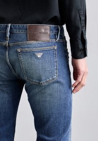 Blå denimjeans med Emporio Armani-läderetikett och logotyp på bakfickan, bärs tillsammans med en svart långärmad skjorta.