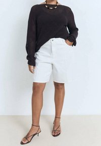 Haut noir en maille à manches longues avec une coupe ample, porté avec un short en denim blanc et des talons noirs à lanières. Accessoires remarquables : un collier épais.