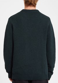 Persoon in een donkergroen gebreid sweater met lange mouwen, zichtbaar van achteren tegen een effen witte achtergrond.