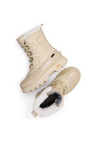Bottes imperméables beige avec une texture lisse et brillante, col doublé de fourrure et semelle à crampons. Les éléments métalliques notables incluent des œillets hexagonaux et un languette de traction.