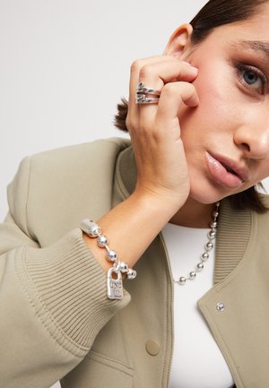 Bracelet en argent avec de grosses perles et un pendentif en forme de cadenas, porté sur un poignet de couleur peau. La veste du modèle est vert olive avec des poignets côtelés.