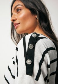 Striped sweater in white and black, featuring a textured knit, round decorative buttons on the shoulder, and ribbed detailing along the neckline.