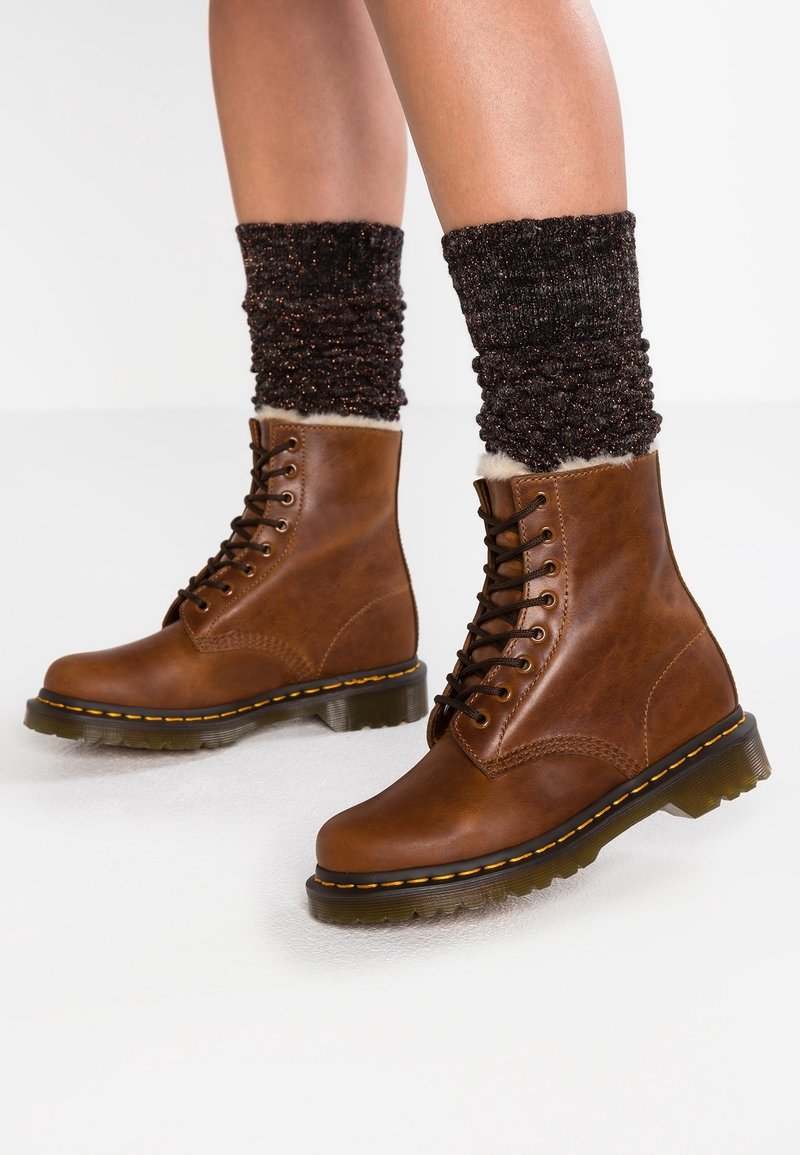 Jambes portant des bottes montantes en cuir marron avec des chaussettes épaisses noires retroussées au-dessus des chevilles sur un sol blanc.