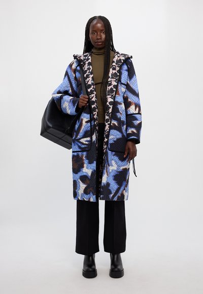 Κουβερτίνα μπλε και μαύρου σχεδίου παλτό με κουκούλα, με μαύρες λεπτομέρειες και τσέπες, συνδυασμένο με καφέ πουλόβερ και μαύρο παντελόνι.