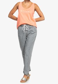 Camiseta sin mangas naranja con logo metálico, pantalones grises de tejido ligero con cordón y sandalias plateadas. Conjunto casual con un ajuste relajado.