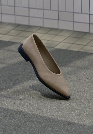 Beige värinen, kärjestään terävä, mustapohjainen tasapohjainen kenkä leijuu harmaan asfaltin yllä lähellä laatoitettua seinää.