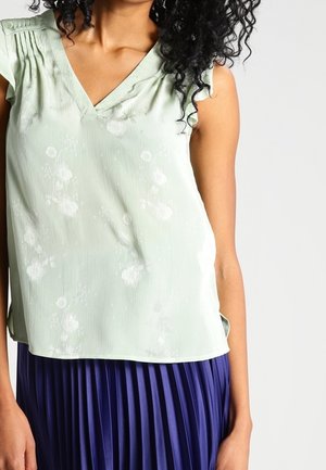 Mujer con una blusa sin mangas de cuello en V de flores verde claro combinada con una falda plisada azul oscuro, de pie contra un fondo liso.