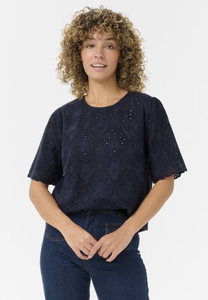 Blouse en coton bleu marine avec broderie œillets, manches courtes bouffantes, encolure ronde et design court. Associée à un jean en denim foncé.