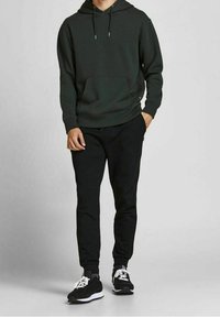 Donkergroene hoodie met een voorkantzak, gecombineerd met zwarte tapse broek en zwarte sportschoenen met witte accenten.