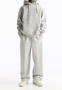 Person iført en lys grå hættetrøje og matchende sweatpants med hvide sneakers, stående med hænderne i lommerne mod en hvid baggrund.