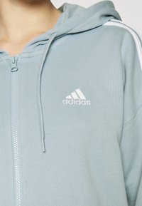 Světlá modrá mikina na zip z texturované látky, s bílým logem na hrudi a bílými pruhy podél ramen.