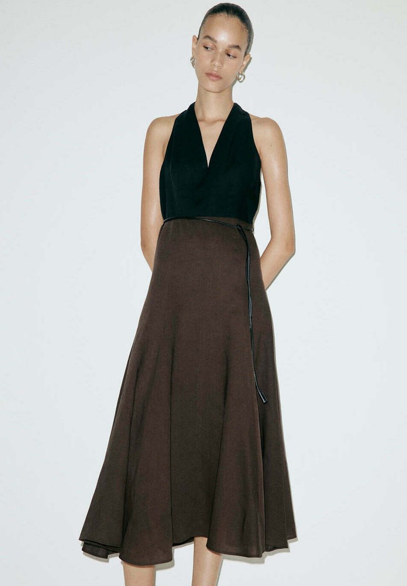 Jeune femme portant un haut noir sans manches à col halter et une jupe midi marron taille haute avec une fine ceinture noire, debout devant un fond blanc.