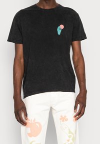 Svart bomullströja med rund nacke och korta ärmar, med en turkos handgrafik som rör vid en rosa cirkel på bröstet.