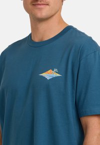 Mann trägt ein blaues T-Shirt mit einem kleinen bunten Strandmotiv, das eine Sonne, Wellen und eine Palme auf der linken Brust zeigt.