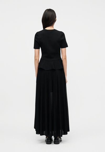 Γυναίκα με μακριά σκούρα μαλλιά, φορώντας μαύρο τοπ με κοντά μανίκια και πεπλούμ, διαφανή μαύρη μακριά φούστα και μαύρα παπούτσια, όρθια με πλάτη προς τον θεατή σε λευκό φόντο.