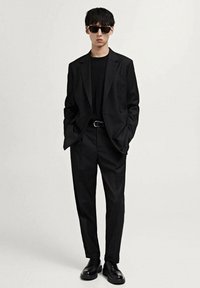 Black tailored suit with a double-breasted jacket and slim-fit trousers. Worn over a black shirt, complemented by black sunglasses and shoes.