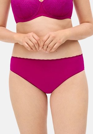 Mujer con sujetador de encaje rosa brillante a juego y bragas lisas, con las manos apoyadas cerca del abdomen inferior sobre un fondo liso.