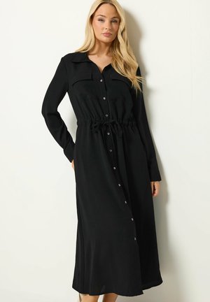 Frau mit langen blonden Haaren trägt ein schwarzes Midi-Kleid mit langen Ärmeln, Knopfleiste und Kordelzug in der Taille und steht vor einer hellen, schlichten Wand.