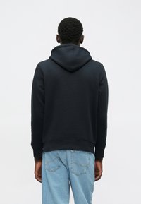 Cilvēks stāv pretī, ar muguru pret skatītājam, ģērbies melnā kapucē un gaiši zilās biksēs uz vienkārša baltā fona.