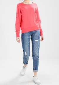 Felpa corallo con triangoli ricamati tono su tono, abbinata a jeans chiari strappati. Sneakers bianche completano l'outfit.