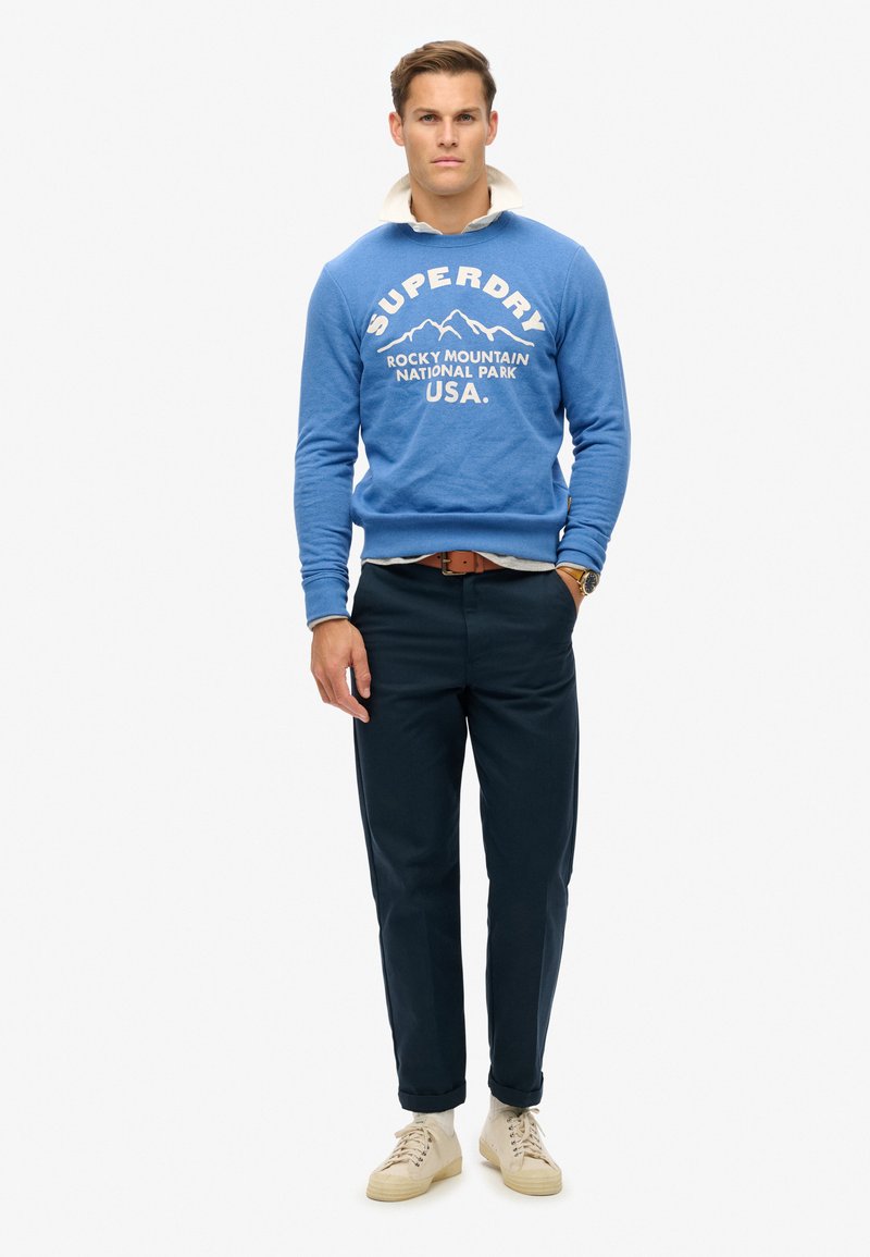Sudadera azul con gráfico de montaña blanca y texto "SUPERDRY", combinada con pantalones oscuros y zapatos beige. El modelo está de pie frente a un fondo blanco.