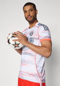 Maglia bianca con accenti rossi, strisce orizzontali grigie e logo del club sul petto. Il modello tiene un pallone da calcio bianco e nero. Pantaloncini rossi visibili.