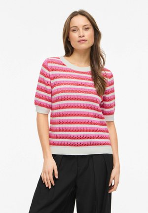 Femme aux longs cheveux portant un haut tricoté rayé rose et rouge à manches courtes et un pantalon noir à jambes larges, debout devant un fond blanc.