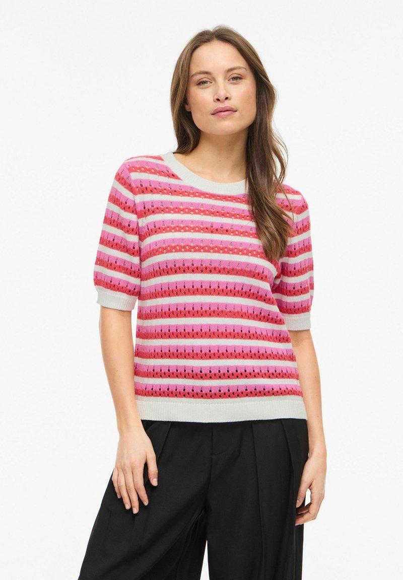 Femme aux longs cheveux portant un haut tricoté rayé rose et rouge à manches courtes et un pantalon noir à jambes larges, debout devant un fond blanc.