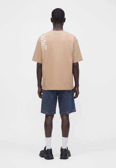Homme portant un t-shirt beige avec un texte blanc sur le haut à gauche dans le dos, un short en jean, des chaussettes blanches et des baskets noires, debout, dos tourné.