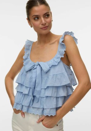 Kvinde iført en lyseblå ærmeløs top med flæser og hulmønster-detaljer, bundet med en sløjfe ved halsudskæringen.
