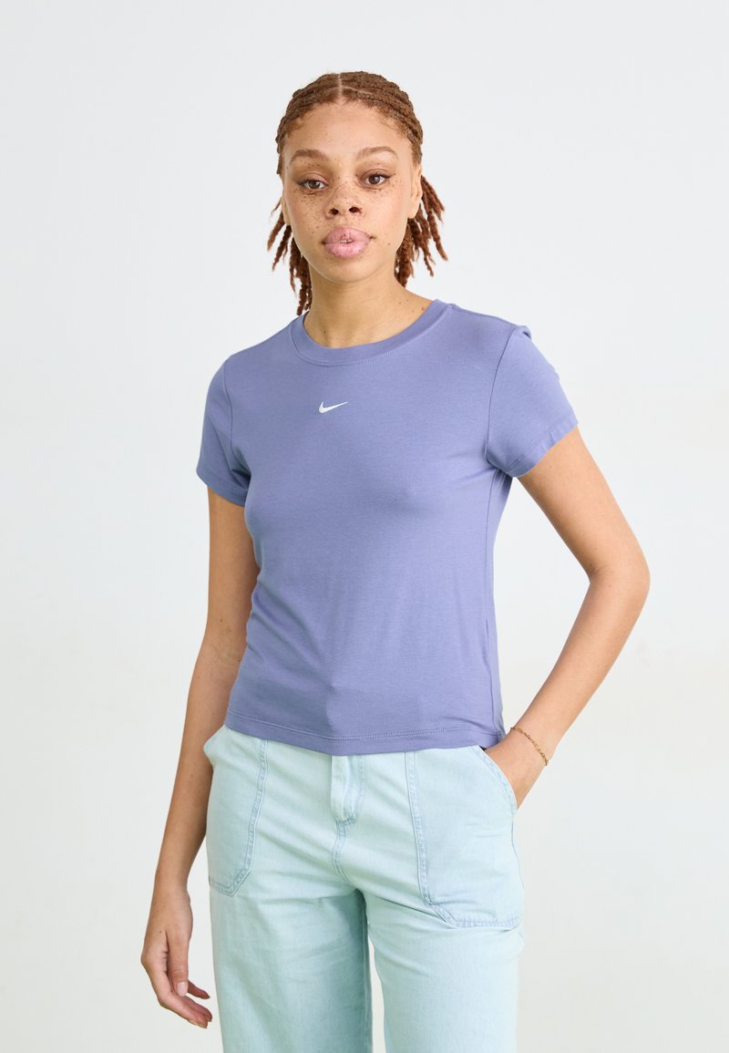 Moteris su pintomis plaukais, vilkinti levandų spalvos Nike marškinėlius ir šviesiai mėlynas kelnes, stovi su viena ranka kišenėje prieš neutralų foną.