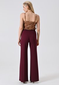 Donna di spalle che indossa un top in satin bronzo con paillettes e pantaloni a vita alta e gamba larga color bordeaux, abbinati a tacchi neri, su uno sfondo neutro.