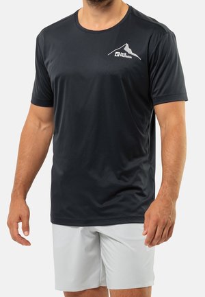 Mężczyzna w czarnej koszulce Jack Wolfskin i jasnoszarych szortach, stojący z ramionami swobodnie opuszczonymi wzdłuż ciała.