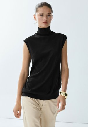 Donna con i capelli raccolti indossa un top nero senza maniche a collo alto, pantaloni beige e un grande braccialetto d'oro al polso destro.